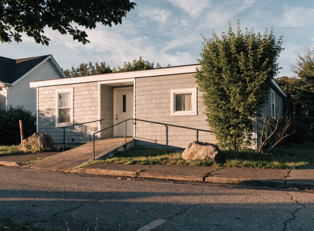 Exterior View Of A Small Single-Story Home Designed For Aging - Small Homes USA Newsletter Front view of a small single-story house with a concrete ramp and black metal handrails, in a sunny suburban street.