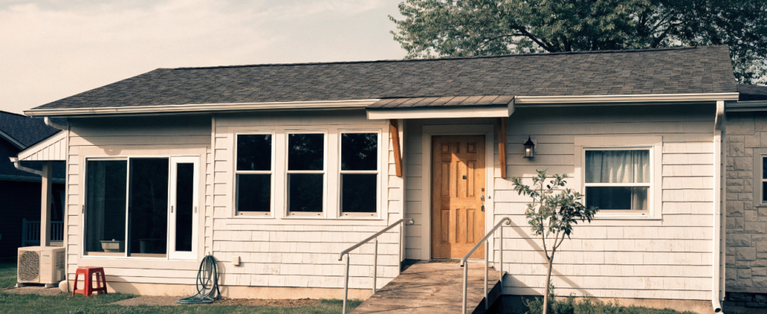 Small House Plans for Aging in Place - Small Homes USA Newsletter Front view of a single-story house with a wooden ramp to the front door, windows along the facade, and a green lawn under a tree behind.